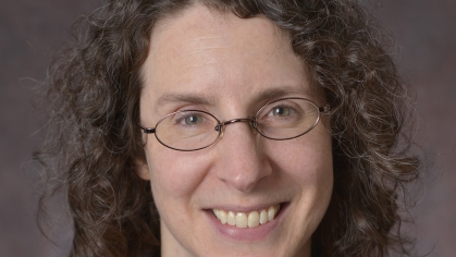 Headshot of smiling woman with glasses and shoulder length brown curly hair wearing a red shirt and black suit jacket