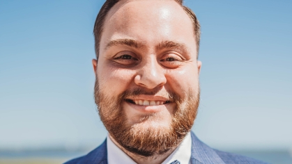 Head shot of male with short light brown hiar, a beard and moustache wearing a blue suit jacket, white button down shirt and a blue patterned bow tie.