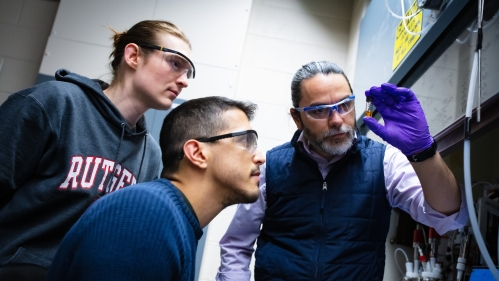male professor wearing dark blue vest and goggles while holding a vial as two male students look on