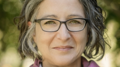 Head shot of woman with short black and grey hair wearing eyeglasses, pink sweater, white shirt, and pink scarf.