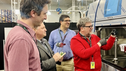 Group of men in a lab setting stand around one man operating a device.