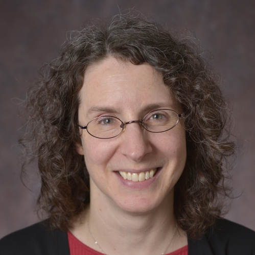 Headshot of smiling woman with glasses and shoulder length brown curly hair wearing a red shirt and black suit jacket
