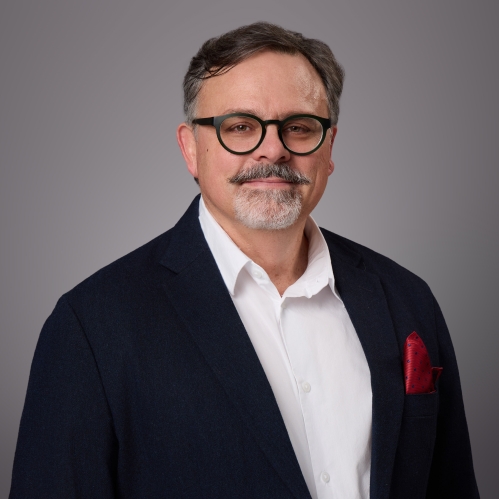 Headshot of male with short hair, grey beard and moustache and eyeglasses wearing a white button down shirt, dark blue suit jacket with a burgundy lapel handkerchief.