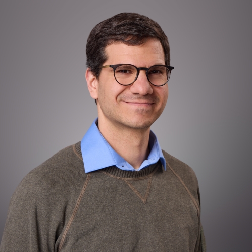 Headshot of male with short brown hair and eyeglasses wearing a blue collared short underneath a khaki pullover sweater.