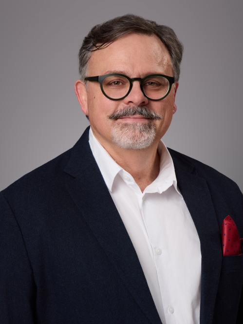 Headshot of male with short hair, grey beard and moustache and eyeglasses wearing a white button down shirt, dark blue suit jacket with a burgundy lapel handkerchief.