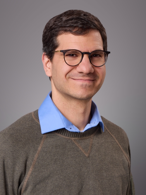 Headshot of male with short brown hair and eyeglasses wearing a blue collared short underneath a khaki pullover sweater.
