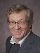 headshot of smiling male wearing glasses, a dark suit and tie with a sweater anAlexander Neimark Headshot