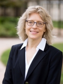 Woman with shoulder lenght blond hair and glasses wearing black suit with white blouse.