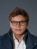 Head shot of Indian male wearing eyeglasses, a white button down shirt and a dark blue jacket.