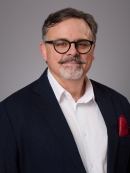 Headshot of male with short hair, grey beard and moustache and eyeglasses wearing a white button down shirt, dark blue suit jacket with a burgundy lapel handkerchief. 