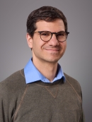 Headshot of male with short brown hair and eyeglasses wearing a blue collared short underneath a khaki pullover sweater.