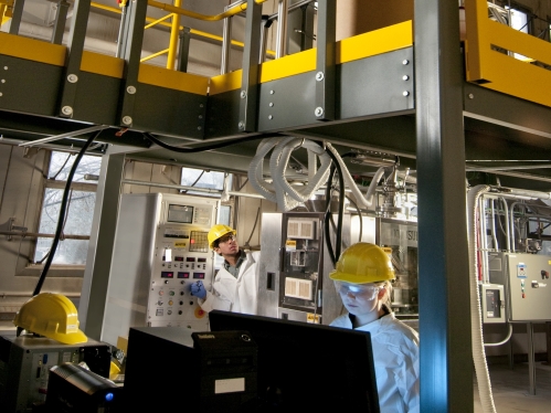 Male and female students in multilevel manufacturing lab wearing yellow hard hats and lab coats.