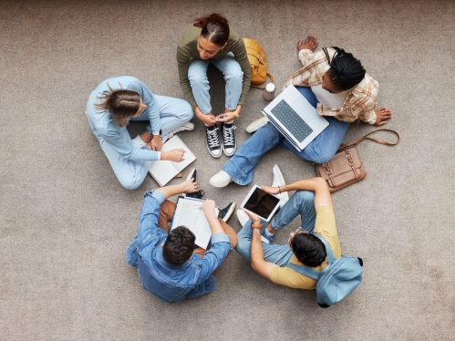 photo looks down from above over five students sitting in a circle on the floor with laptops and backpacks.