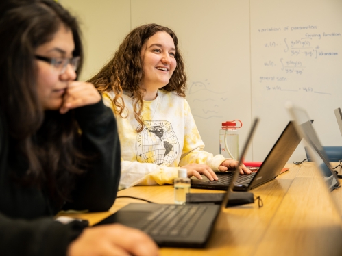 Two female students sitting at table with laptops. One has dark hair and black sweatshirt, the other has brown curly hair and a yellow T-shirt.