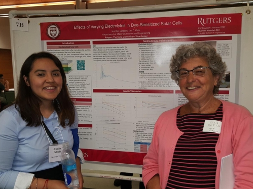 A younger white woman with black hair and light blue shirt stands in front of a research poster along with an older woman with short gray hair and glasses wearing a pink sweater. Both are smiling.