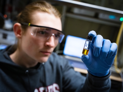 male student in lab wearing goggles looking closely at vile with yellow substance