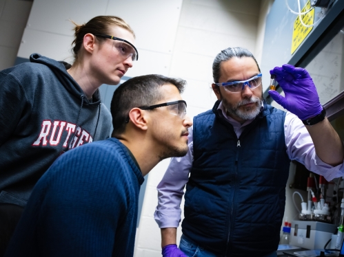 male professor wearing dark blue vest and goggles while holding a vial as two male students look on