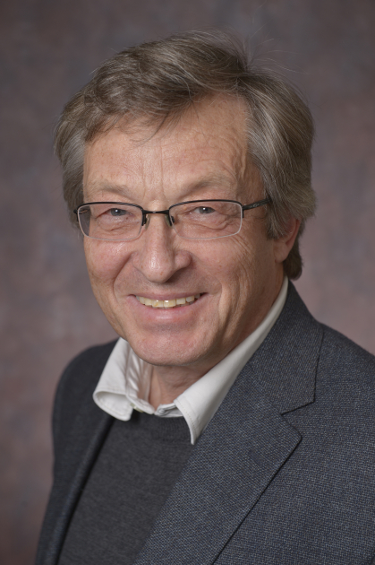headshot of smiling male wearing glasses, a dark suit and tie with a sweater anAlexander Neimark Headshot