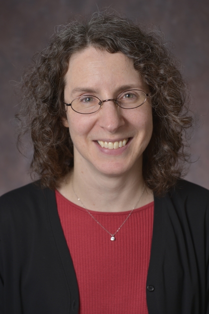 Headshot of smiling woman with glasses and shoulder length brown curly hair wearing a red shirt and black suit jacket