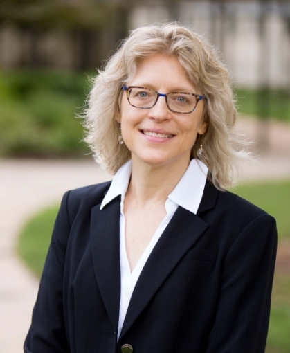 Woman with shoulder lenght blond hair and glasses wearing black suit with white blouse.