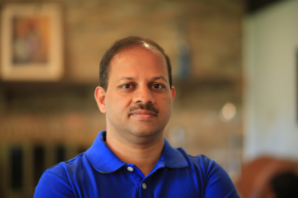 Head shot of Indian male with a moustache and wearing a blue button down shirt.