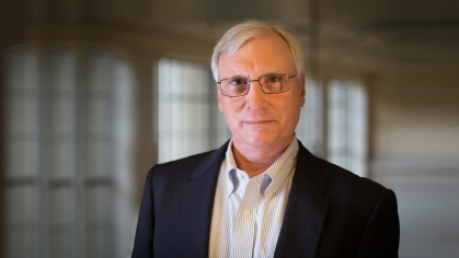 Head shot of male with grey hair with eyeglasses wearing a black suit jacket with a white button down shirt.