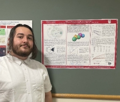 Male student with long brown hair and a bear, wearing a button down white shirt stands next to a poster pinned to a bulletin board.