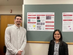 Tall male on the left with a beard weraing a button down shirt and a short female on the right standing  next to their senior design poster which is pinned to a bulletin board.