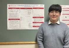 Asian woman with short black hair and eyeglasses stands next to her senior design poster pinned to a bulletin board.