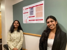 Two female students flank their senior design poster pinned to a bulletin board.