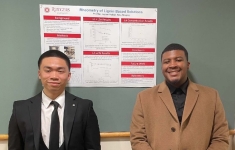Asian male stands on left and Black male on the right flanking their senior design poster pinned to a bulleltin board.