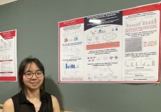Female student with short black hair and eyeglasses stands next to her senior design which is pinned to a bulletin board.poster 